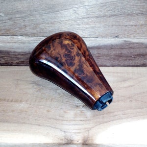 May include: A brown wooden gear shift knob with a dark brown wood grain pattern. The knob has a black plastic base with a metal clip.