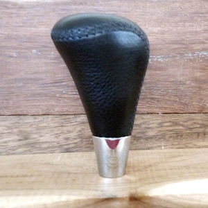 以下が含まれることがあります： 銀色のベースが付いた黒い革製のシフトノブ。ノブにはステッチ模様とテクスチャ加工が施されています。