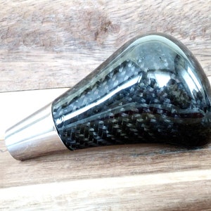 May include: A black carbon fibre gear shift knob with a silver metal base.