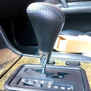 Może przedstawiać: Czarny skórzany gałka zmiany biegów z chromowanym pierścieniem wokół podstawy. Drążek zmiany biegów znajduje się w pozycji neutralnej. Schemat zmiany biegów jest widoczny z numerami 1, 2, 3 i 4D.