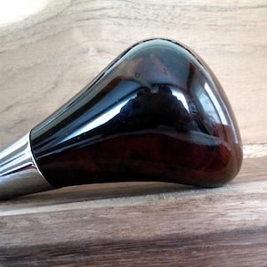 Może przedstawiać: Gałka zmiany biegów z ciemnego drewna z chromowaną podstawą. Gałka ma błyszczące wykończenie.