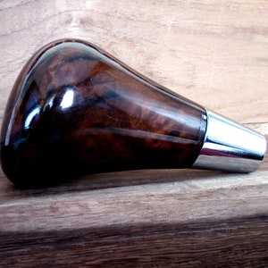 Może przedstawiać: Gałka zmiany biegów z ciemnego drewna z chromowanym akcentem. Gałka ma unikalny wzór usłojenia drewna.