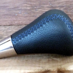 Puede incluir: Pomo de cambio de marchas de cuero negro con costuras azules y una base de metal plateado.