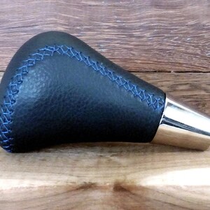 以下が含まれることがあります： 青いステッチとシルバーの金属製のベースが付いた、黒い革製のシフトノブです。
