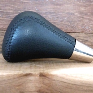 Puede incluir: Pomo de cambio de marchas de cuero negro con base de metal plateado. El pomo tiene una costura cosida alrededor de la parte superior.