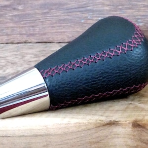 Puede incluir: Pomo de palanca de cambios de cuero negro con base plateada y costuras rosas.