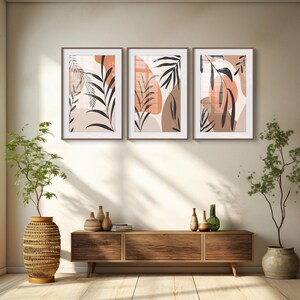 May include: Three framed abstract art prints with a minimalist design featuring brown and orange leaves and shapes on a white background. The prints are hanging on a beige wall above a wooden console table.