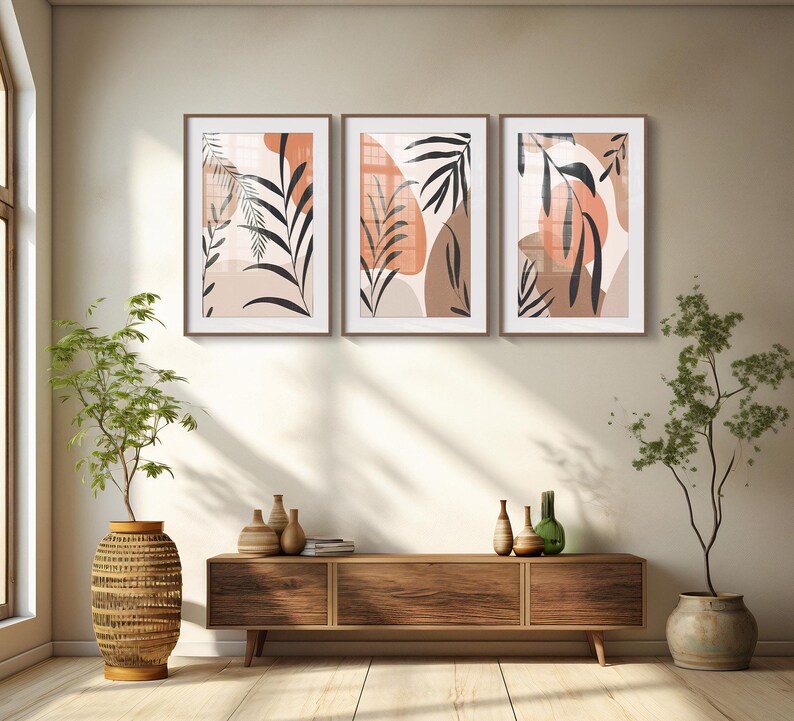 May include: Three framed abstract art prints with a minimalist design featuring brown and orange leaves and shapes on a white background. The prints are hanging on a beige wall above a wooden console table.