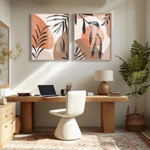 May include: Two abstract art prints with brown and orange geometric shapes and black leaf silhouettes. The prints are framed in a light wood frame and are hanging on a light beige wall above a wooden desk.