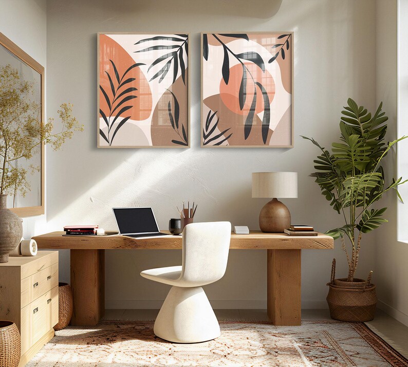 May include: Two abstract art prints with brown and orange geometric shapes and black leaf silhouettes. The prints are framed in a light wood frame and are hanging on a light beige wall above a wooden desk.