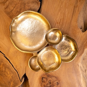May include: A collection of five irregularly shaped, gold-colored metal bowls. The bowls vary in size and are arranged together on a wooden surface with a natural grain pattern. The bowls have a hammered texture.