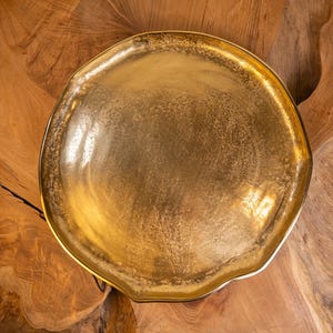 May include: A round, shallow metal tray with a metallic gold finish. The surface of the tray features a textured, mottled appearance with variations in light and shadow across the circular form, creating a reflective and uneven golden sheen.