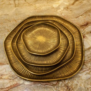 May include: A set of four nested, organically shaped, gold-colored serving trays. Each tray has a textured, ridged surface radiating from the center. The trays are arranged on a marble-patterned surface.