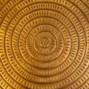 May include: Close-up of a golden, textured surface with a concentric circular pattern. The design features a series of etched lines and shapes radiating outwards from a central point, creating a detailed and ornate appearance. The overall effect is warm and inviting.