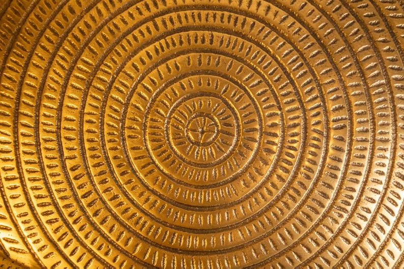 May include: Close-up of a golden, textured surface with a concentric circular pattern. The design features a series of etched lines and shapes radiating outwards from a central point, creating a detailed and ornate appearance. The overall effect is warm and inviting.