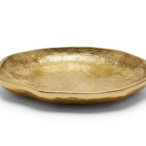 May include: A shallow, circular tray with a metallic gold finish and a textured, hammered surface. The edges of the tray are slightly irregular and organic in shape, creating a subtle, uneven rim that contrasts with the smooth, polished base.