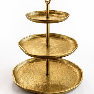 May include: A three-tiered serving stand with a gold finish. The stand has three round trays of varying sizes, connected by a central pole with a ring handle at the top. The trays have a textured surface.
