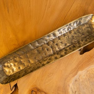 May include: A rectangular, antique brass tray with a textured surface. The tray has rounded corners and is set against a warm, wooden background. This decorative tray is ideal for serving or displaying items.