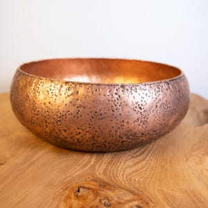 May include: A copper-colored bowl with a textured surface. The bowl has a rounded shape and sits on a wooden surface. The interior of the bowl has a smooth, reflective surface. The bowl is a decorative item.