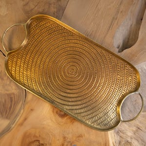 May include: A rectangular gold-colored metal tray with a textured, concentric circle design. The tray has rounded edges and two circular handles. The tray is displayed on a wooden surface.