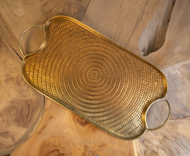 May include: A rectangular gold-colored metal tray with a textured, concentric circle design. The tray has rounded edges and two circular handles. The tray is displayed on a wooden surface.