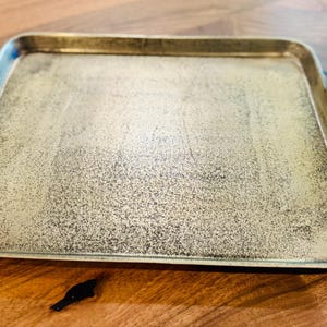 May include: A rectangular metal tray with a distressed, speckled finish. The tray has branch-shaped handles on each side. The color is a mix of gold and silver, with a slightly textured surface. A decorative serving piece.
