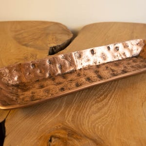 May include: A rectangular copper tray with a hammered texture. The tray has rounded corners and is displayed on a wooden surface. The copper has a warm, reddish-brown color.