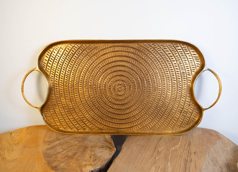 May include: A rectangular gold-colored tray with a circular textured design and two rounded handles. The tray is made of metal and sits on a wooden surface. The tray's design features concentric circles.