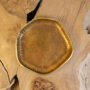 May include: A decorative, gold-colored tray with a unique, organic shape. The tray has a textured, sunburst-like pattern radiating from the center. The tray is displayed on a light brown wooden surface.