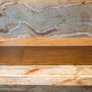 May include: A rectangular, gold-toned metal tray with raised sides and handles. The tray has a textured surface and is set against a marble background with shades of brown, beige, and white. The tray is approximately 24 inches long.