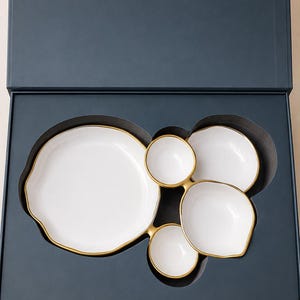 May include: A set of five white ceramic bowls with gold-painted rims, arranged in a dark blue presentation box. The box lid features the gold text BOWL & FRAME and The Luxury of Together. The bowls vary in size and shape, nested in a custom cutout.