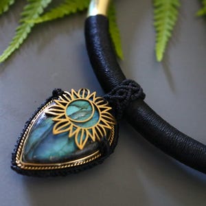 May include: A handcrafted necklace featuring a teardrop-shaped pendant with a gold sunflower design and a blue-green stone. The pendant is attached to a black cord necklace with gold accents. The necklace is displayed on a gray surface with green foliage.