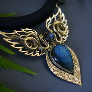 May include: A gold-toned pendant necklace with a large, teardrop-shaped blue gemstone. The pendant features intricate snake and wing designs, with a black cord necklace. The background is a dark blue with a fern leaf.