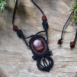 Peut inclure: Collier macramé fait à la main avec une grosse perle ovale en bois, rayée marron et beige. La perle est sertie dans un motif macramé noir, avec des détails rouge-brun. Le collier a des cordons noirs réglables. Présenté sur une surface en bois.