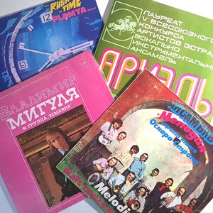 May include: A collection of vintage vinyl records with colorful album covers. The covers feature text and images, including a clock design, a portrait, and group photos. The records are arranged on a white surface.