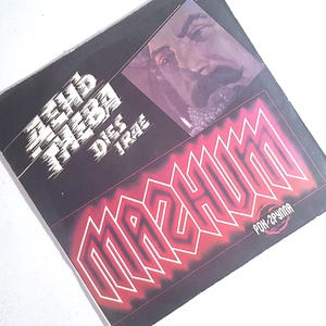 Può includere: Una copertina quadrata per disco in vinile con sfondo nero e un'illustrazione stilizzata di un volto nell'angolo in alto a destra. Il testo recita "ДЕНЬ ГНЕВА DIES IRAE" in bianco e "MAGNUM" in grandi lettere rosse luminose.