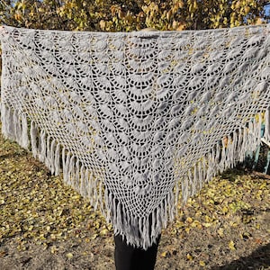 Könnte beinhalten: Ein grauer Häkel-Schal mit Fransenbordüre. Der Schal hat ein kreisförmiges Muster mit einem gewellten Rand.