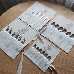 Può includere: Un set di quattro rotoli di posate in lino, ciascuno contenente posate. I rotoli sono beige con un motivo a foglie e accenti di foglie ricamate. Ogni rotolo ha un legaccio in tessuto. Forchette, coltelli e cucchiai sono visibili.