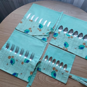 May include: A set of four teal fabric utensil holders with floral patterns. Each holder contains stainless steel silverware: knives, forks, and spoons. The holders are arranged on a wooden table.