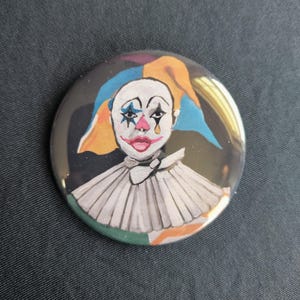 Puede incluir: Un botón redondo con la cara de un payaso pintada. El payaso tiene pintura facial blanca, una nariz rosa y labios rojos. El pelo del payaso es una mezcla de azul, naranja y amarillo. El payaso lleva un cuello blanco con volantes.