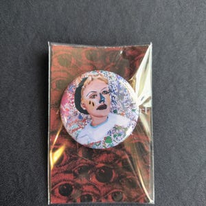 Puede incluir: Un botón redondo con un retrato de una persona con maquillaje oscuro y un fondo floral. El pin está empaquetado en una tarjeta roja con un patrón de ojos.