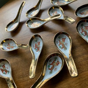 May include: A collection of white ceramic soup spoons featuring gold-toned rims and handles. Each spoon is decorated with a detailed orange and green dragon motif centered in the bowl. The spoons are arranged in a scattered pattern.
