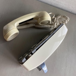 May include: An off-white vintage telephone with a handset and coiled cord. The base of the phone is angled with a silver metal cradle. The handset has a speaker and a microphone.