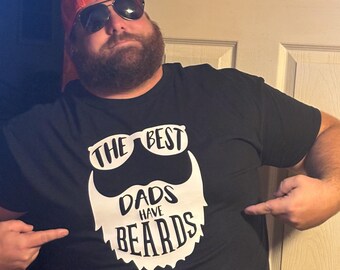 Puede incluir: Camiseta negra con un gráfico blanco de una barba con forma de gafas de sol y el texto "THE BEST DADS HAVE BEARDS". La persona de la imagen lleva una gorra naranja y gafas de sol.