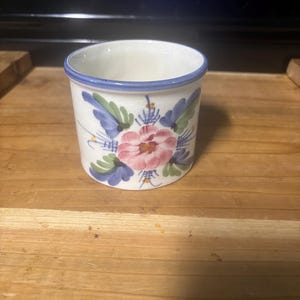 May include: A small, cylindrical ceramic cup with a white interior and a blue rim. The exterior is decorated with a floral design featuring pink, blue, and green elements. The cup is likely used for drinking or as a decorative item.