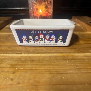 May include: A white rectangular ceramic dish with a blue border featuring cartoon snowmen and the text "LET IT SNOW." The dish is on a wooden surface, with a lit candle in a decorative jar in the background.