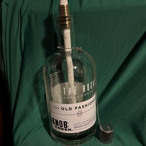 May include: A clear glass bottle repurposed as an oil lamp. It features a brass fitting, a white wick, and a metal cap attached by a chain. The bottle has a label that reads "ON THE ROCKS" and "THE OLD FASHIONED".