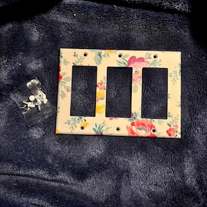 May include: A decorative triple light switch cover with a floral pattern. The cover features a cream background with colorful flowers in shades of pink, yellow, and red, with green leaves. Mounting screws are included.