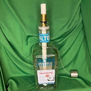 May include: A repurposed Altos Margarita bottle transformed into a decorative oil lamp. The clear glass bottle features blue and white labels with the text "ALTOS MARGARITA" and "Strawberry." A wick and metal hardware are attached to the bottle.