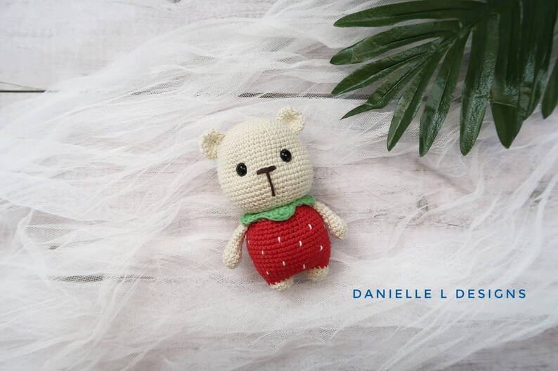 May include: A crocheted stuffed toy bear wearing a red and green strawberry costume. The bear is sitting on a white fabric background.